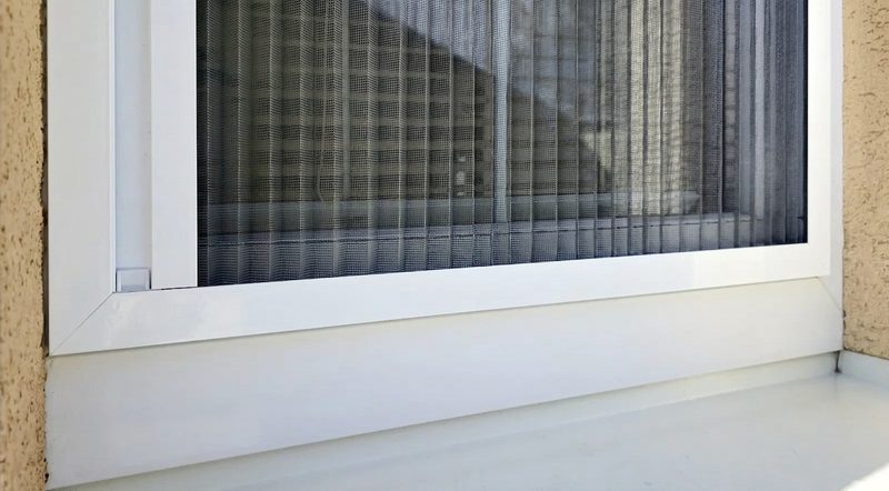 Spodní hrana bílého plastového okna s plisovanou sítí a drobným úchytem na rámu
