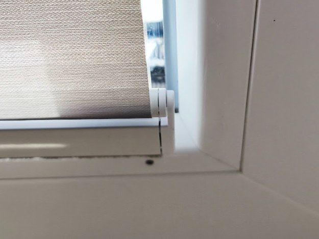 Spodní roh plastového okna, pod spodní lištou nové látkové rolety díra po vrutu v rámu