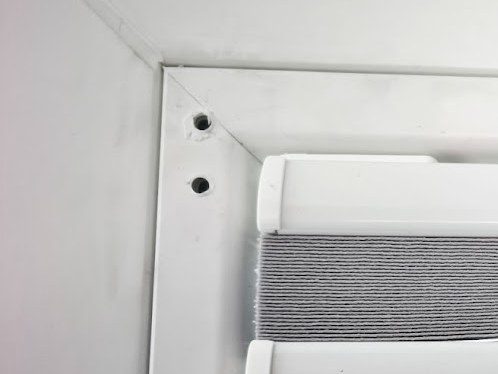 Horní roh bílého plastového okna se dvěma dírami po vrutech staré žaluzie v rámu, vedle nich horní profil nového plisé